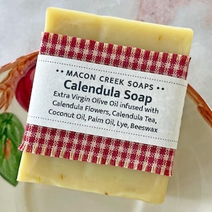 May include: A bar of Calendula Soap wrapped in red and white checkered fabric. The label reads "MACON CREEK SOAPS... Calendula Soap Extra Virgin Olive Oil infused with Calendula Flowers, Calendula Tea, Coconut Oil, Palm Oil, Lye, Beeswax"
