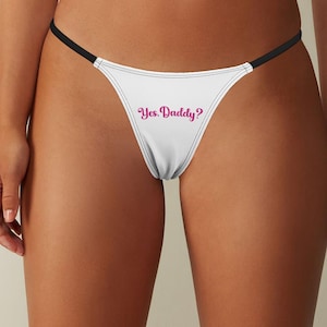 Könnte beinhalten: Weißer String-Tanga mit schwarzen Trägern. Auf der Vorderseite des Tangas steht der Text "Yes, Daddy?" in rosa Schrift. Die Unterwäsche ist aus weichem Material und hat ein minimalistisches Design.
