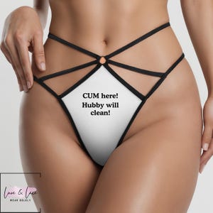 May include: White and black lingerie with the text "CUM here! Hubby will clean!" The lingerie features a diamond-shaped front panel and crisscross straps. The lingerie is from the brand Love & Lace.
