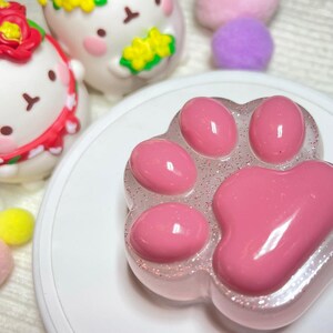 May include: A pink paw-shaped soap with a glittery, clear border sits on a white plate. The soap has five rounded pads. In the background are two white bunny figurines with floral accents and colorful pom-poms.