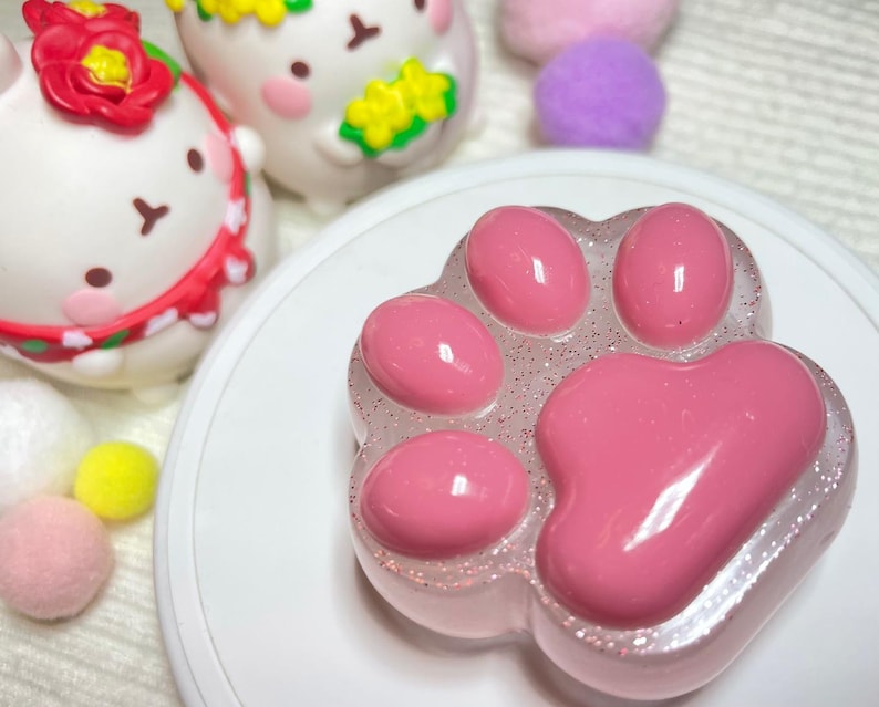 May include: A pink paw-shaped soap with a glittery, clear border sits on a white plate. The soap has five rounded pads. In the background are two white bunny figurines with floral accents and colorful pom-poms.