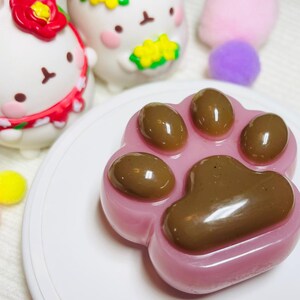 May include: A pink, paw-shaped dessert with brown paw pads, presented on a white plate. The image also features several white, cartoon-like figures with red and yellow accents, and colorful pom-poms.