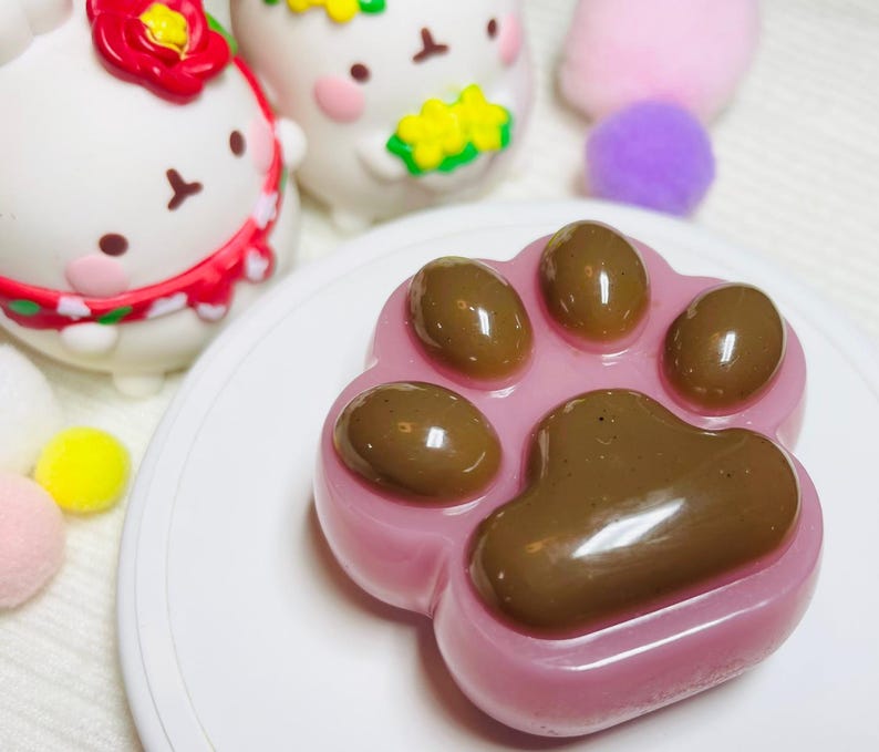 May include: A pink, paw-shaped dessert with brown paw pads, presented on a white plate. The image also features several white, cartoon-like figures with red and yellow accents, and colorful pom-poms.