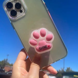 May include: Clear phone case with a pink paw print phone grip. The paw print has the word "SHINE" in white text with a small red heart. The case has a clear back and iridescent edges.
