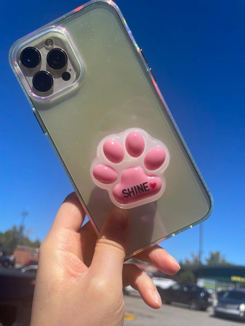 May include: Clear phone case with a pink paw print phone grip. The paw print has the word "SHINE" in white text with a small red heart. The case has a clear back and iridescent edges.