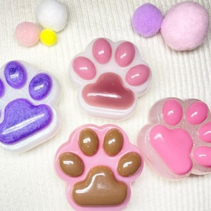 May include: Several resin paw-shaped decorations in shades of pink, purple, and brown, some with glitter. The image also shows small, fluffy pom-poms in white, pink, and yellow.