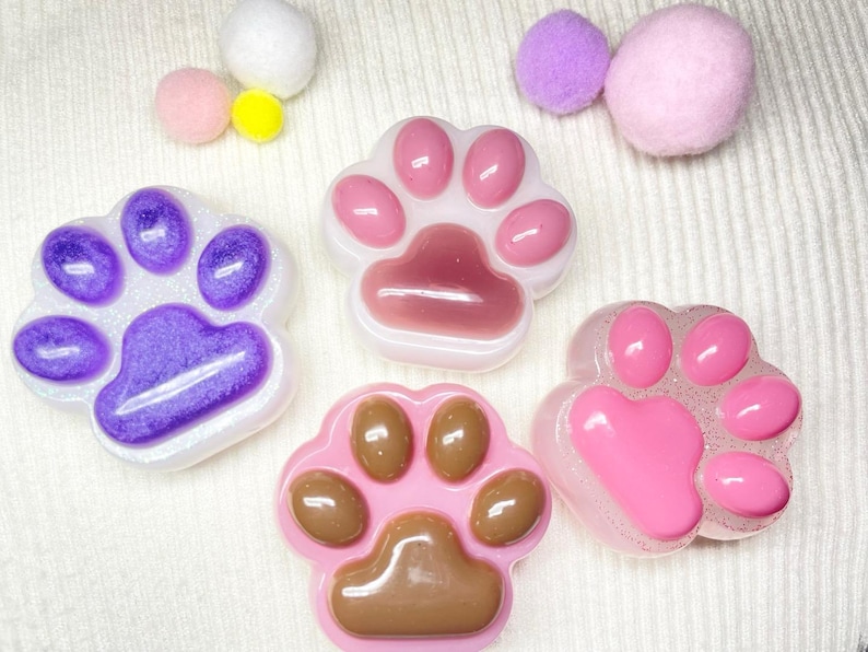 May include: A collection of paw-shaped resin art pieces in various shades of pink, purple, and brown. Some pieces have glitter accents. The image also includes small, fluffy pom-poms in white, pink, and yellow.