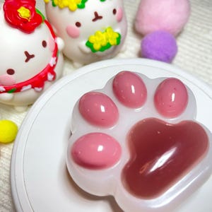 May include: A white paw-shaped soap with pink pads sits on a white plate. Two cartoon figurines and several colorful pom-poms are in the background. The figurines are white with floral accents.