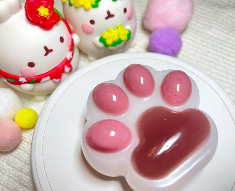 May include: A white paw-shaped soap with pink pads sits on a white plate. Two cartoon figurines and several colorful pom-poms are in the background. The figurines are white with floral accents.