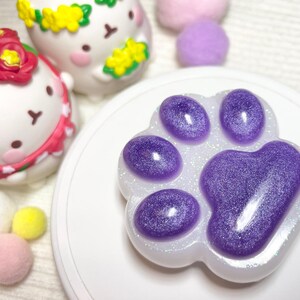 May include: A paw-shaped resin item with a glittery purple design. The paw print is surrounded by a white border with glitter. In the background are two white figurines with floral headbands and several small, colorful pom-poms.
