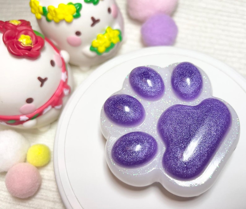 May include: A paw-shaped resin item with a glittery purple design. The paw print is surrounded by a white border with glitter. In the background are two white figurines with floral headbands and several small, colorful pom-poms.