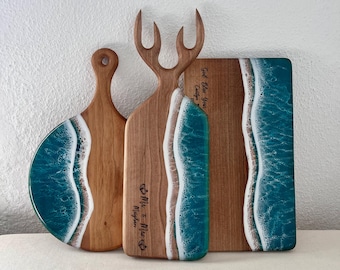 Könnte beinhalten: Drei Holzschneidebretter mit Strandmotiv. Die Bretter zeigen türkisfarbenes Harz, das den Ozean darstellt, mit weißem Harz für die Wellen. Eines hat einen Griff, ein anderes eine geweihartige Form, und das dritte ist rechteckig.