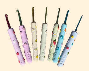 May include: A collection of seven crochet hooks with colorful handles. Each handle is decorated with small, raised floral and fruit designs. The hooks are in shades of pink, green, yellow, and blue. The sizes are marked on the handles.