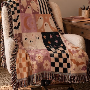 Könnte beinhalten: Eine gewebte Wohndecke mit Patchwork-Design, das Geister, Fledermäuse, Sterne, Blumen und Karomuster in Orange-, Rosa-, Schwarz- und Cremetönen zeigt. Die Decke hat einen Fransenrand und liegt über einem weißen, strukturierten Bürostuhl.