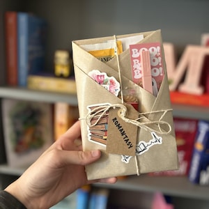 May include: A brown paper package tied with twine and decorated with stickers. The stickers include a stack of books, a floral design, a pink heart with a book design, and a tag that says "Romantasy".