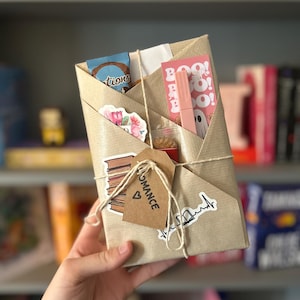 Op de afbeelding: Een bruine papieren verpakking, omwikkeld met touw en versierd met stickers. De verpakking bevat een boek, een klein zakje met lekkernijen, een lipgloss en een klein kaartje met de tekst "Romance".