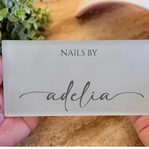 May include: A rectangular, frosted glass sign with the text "NAILS BY" above the name "adelia" in a cursive font. The sign is held by hands, with greenery visible in the background.