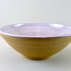 Puede incluir: Un cuenco de cerámica hecho a mano con un diseño ancho y poco profundo. El exterior es de color marrón dorado texturizado, mientras que el borde interior es de un suave lavanda pálido. Adecuado para servir o exhibir.