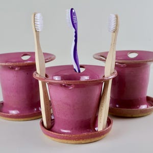 Puede incluir: Tres portacepillos de dientes de cerámica magenta hechos a mano. Cada soporte tiene una base circular y una parte superior ensanchada con un agujero. Dos soportes tienen cepillos de dientes de madera y uno tiene un cepillo de dientes morado.