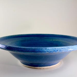 Puede incluir: Un cuenco de cerámica azul hecho a mano con un diseño ancho y poco profundo. El cuenco tiene una superficie texturizada y un borde ligeramente elevado. El interior y el exterior son de un azul vibrante, con un borde azul más claro. El cuenco es adecuado para servir.