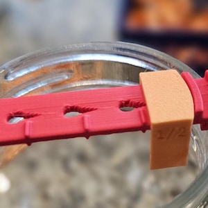 May include: Red plastic jar opening tool with a 1/2 inch measurement on the side. The tool has a series of ridges on one side and holes on the other side.