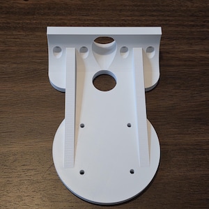 May include: A white, 3D-printed plastic bracket with a circular base and a rectangular top. The bracket has several holes and vertical ribbed supports. The design appears to be for mounting or securing an object.