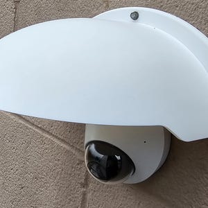 May include: A white security camera with a dome-shaped lens and a protective white cover. The camera is mounted on a textured, light brown wall. The lens is black and reflective.