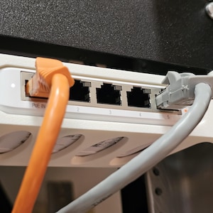 May include: A white network switch with five Ethernet ports, labeled 2 through 5, and an orange and gray Ethernet cable plugged in. The switch is mounted in a white bracket and attached to a black and silver metal structure.