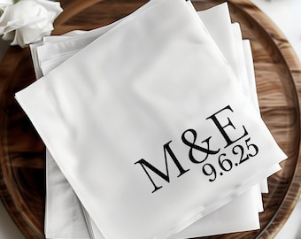 May include: A stack of square white paper napkins featuring black printed text that reads M&E followed by the date 9.6.25. The napkins are folded and layered, displaying a clean, smooth surface with sharp, dark lettering centered on the top napkin.