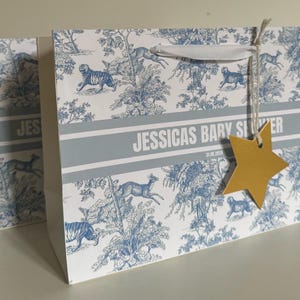 May include: Gift bag with a blue and white toile pattern featuring tigers and deer. The bag has a white ribbon handle and a gold star-shaped tag. The text "JESSICAS BARY SILVER" is printed on the bag.