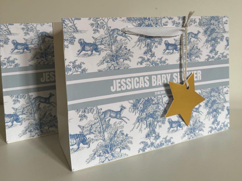 May include: Gift bag with a blue and white toile pattern featuring tigers and deer. The bag has a white ribbon handle and a gold star-shaped tag. The text "JESSICAS BARY SILVER" is printed on the bag.