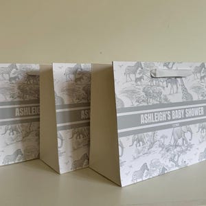 May include: Three gift bags with a safari animal print in gray and white. The bags have a gray band with the text "ASHLEIGH'S BABY SHOWER". Each bag has a white handle.