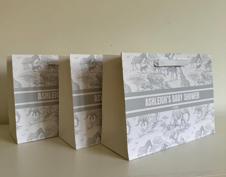 May include: Three gift bags with a safari animal print in gray and white. The bags have a gray band with the text "ASHLEIGH'S BABY SHOWER". Each bag has a white handle.