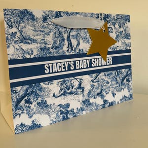 May include: Gift bag with a blue and white toile pattern, featuring the text "STACEY'S BABY SHOWER." A gold star-shaped tag hangs from the handle. The bag has a white handle and a white body.