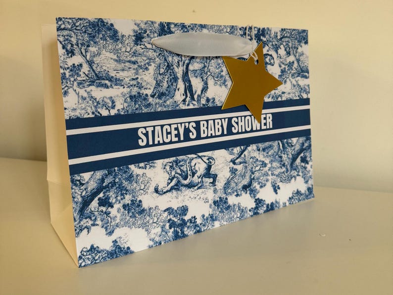 May include: Gift bag with a blue and white toile pattern, featuring the text "STACEY'S BABY SHOWER." A gold star-shaped tag hangs from the handle. The bag has a white handle and a white body.