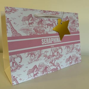 May include: A pink and white gift bag with a safari animal print, including giraffes, zebras, and elephants. The bag has a gold star-shaped tag and the name "SERAPHINA" printed on it.