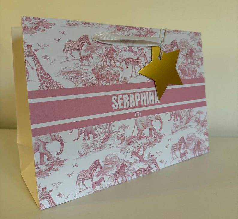 May include: A pink and white gift bag with a safari animal print, including giraffes, zebras, and elephants. The bag has a gold star-shaped tag and the name "SERAPHINA" printed on it.