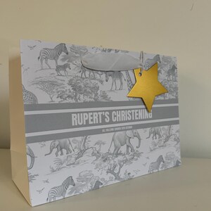 May include: A gift bag with a safari animal print, featuring zebras and elephants. The bag has a gray and white color scheme, with the text "RUPERT'S CHRISTENING" and a gold star-shaped tag attached with a white ribbon.