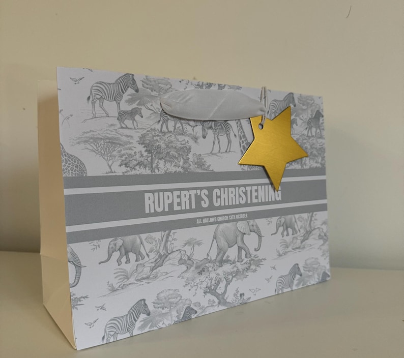 May include: A gift bag with a safari animal print, featuring zebras and elephants. The bag has a gray and white color scheme, with the text "RUPERT'S CHRISTENING" and a gold star-shaped tag attached with a white ribbon.