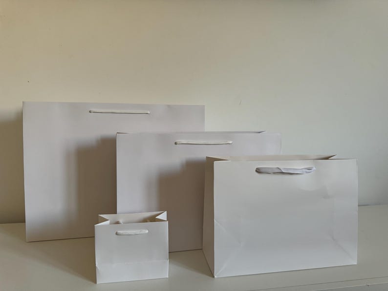 May include: Four white gift bags of varying sizes are arranged on a white surface. Each bag has a matching white handle. The bags are empty and have a glossy finish, suggesting they are made of paper or cardstock.