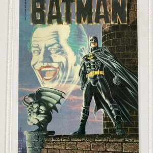 May include: A Batman comic book cover featuring Batman in a black suit with a yellow belt and bat symbol. The Joker's face is in the background. The cover reads "BATMAN" and "THE OFFICIAL COMIC ADAPTATION OF THE WARNER BROS. MOTION PICTURE."