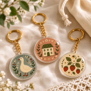 May include: Three circular acrylic keychains with gold-toned metal rings and chains. One features a white goose, one a small house with a green roof, and one a strawberry plant with red berries. Each design is framed by a delicate floral wreath border.