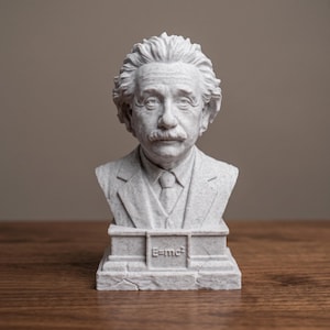 May include: A light gray, speckled bust of Albert Einstein featuring his signature wild hair and mustache. The figure wears a suit jacket, vest, and tie. The base of the sculpture displays the embossed scientific equation E=mc² in a square frame.