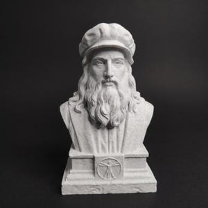 Pode incluir: Um busto escultural cinza-claro de um homem barbudo usando um boné. A figura tem cabelos longos e uma barba detalhada. A base da escultura apresenta um emblema circular representando uma figura humana com braços e pernas estendidos.