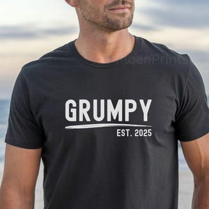 May include: Black crew neck t-shirt with the word "GRUMPY" in white block letters above a white underline and the text "EST. 2025". The shirt is worn by a person on a beach.