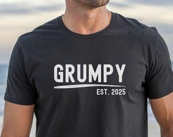 Może przedstawiać: Czarny t-shirt z okrągłym dekoltem z napisem "GRUMPY" białymi literami nad białą linią i tekstem "EST. 2025". Koszulka jest noszona przez osobę na plaży.