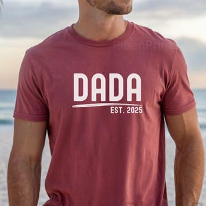 May include: A maroon t-shirt with white text that reads "DADA EST. 2025".