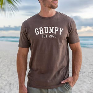 May include: A brown t-shirt with the text "GRUMPY EST. 2025" printed in white.