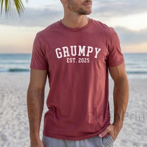 Może przedstawiać: Bordowy t-shirt z białym napisem "GRUMPY EST. 2025". Osoba w t-shircie stoi na plaży.