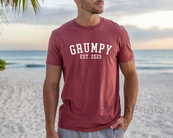 Może przedstawiać: Bordowy t-shirt z białym napisem "GRUMPY EST. 2025". Osoba w t-shircie stoi na plaży.